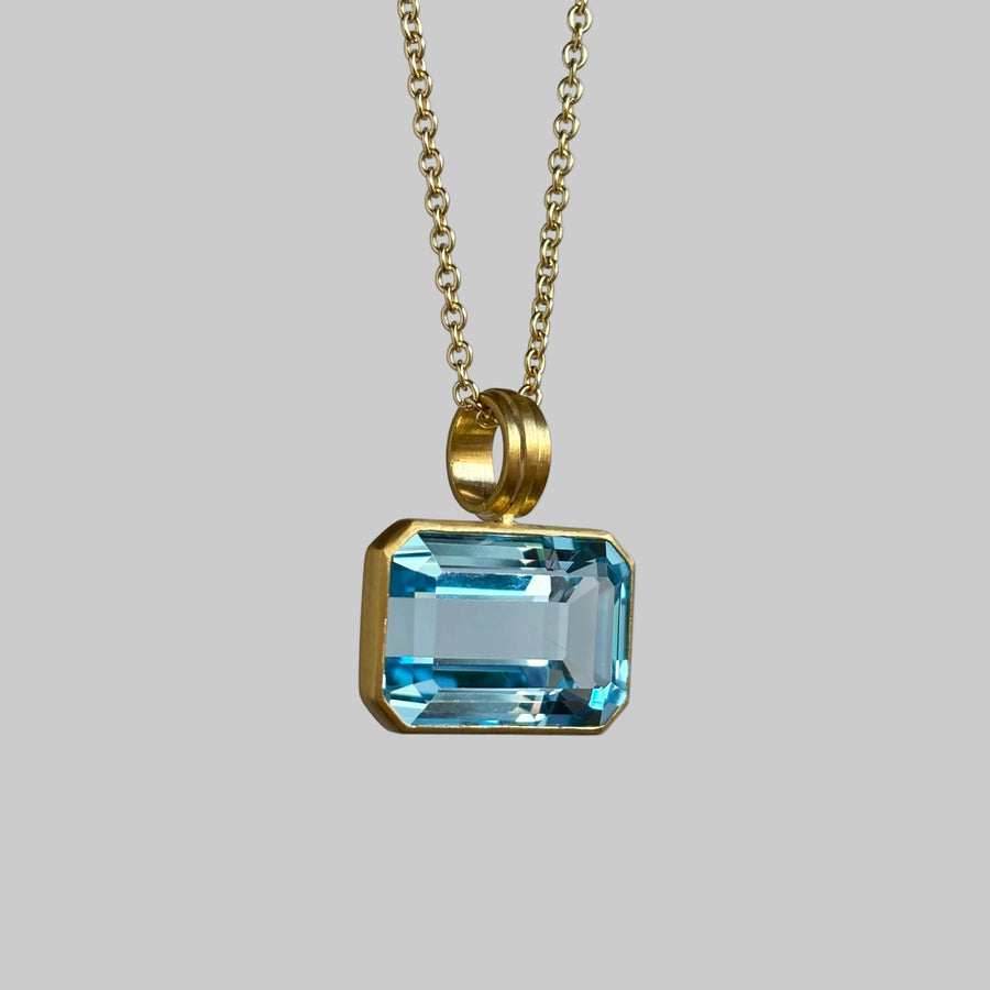 A gold chain necklace with a rectangular light blue gemstone pendant in a gold bezel setting, shown against a neutral light gray background.