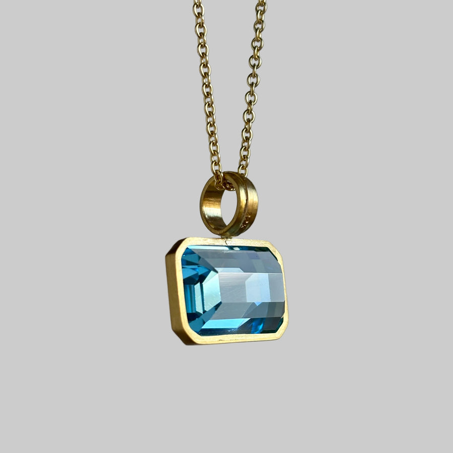 A gold chain necklace with a rectangular light blue gemstone pendant in a gold bezel setting, shown against a neutral light gray background.