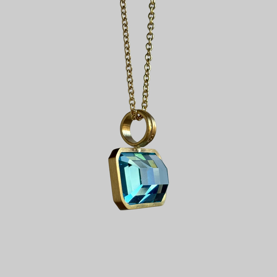 A gold pendant necklace featuring a faceted, beveled rectangular light blue gemstone in a gold bezel setting, suspended from a gold chain against a neutral gray background.