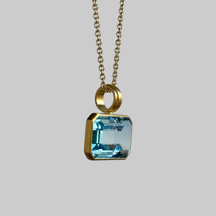 A gold pendant necklace featuring a square-cut light blue gemstone in a gold bezel setting, suspended from a gold chain against a neutral gray background.