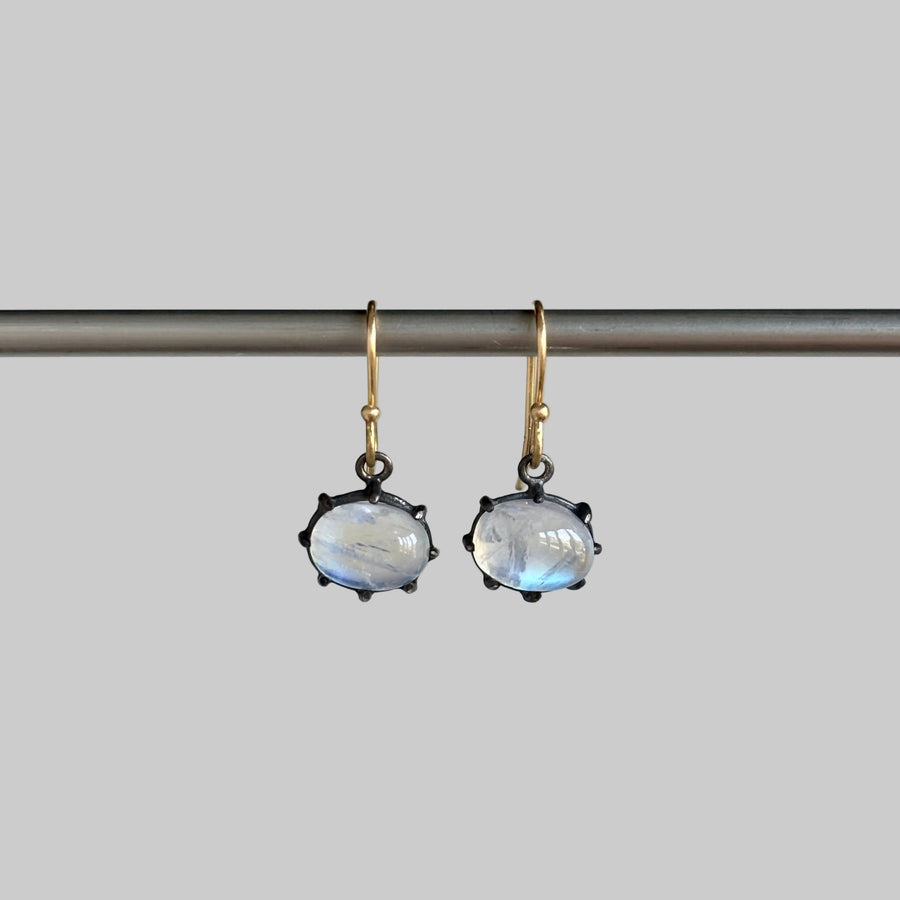 A pair of dangling earrings with oval milky white stones in dark metal settings and gold hooks, hanging from a horizontal bar against a plain light background.