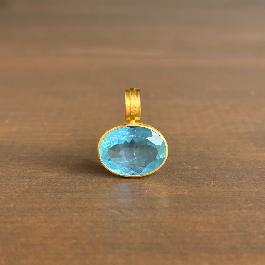 An oval-cut light blue gemstone pendant set in a gold bezel with a matching gold bail, resting on a brown wooden surface.