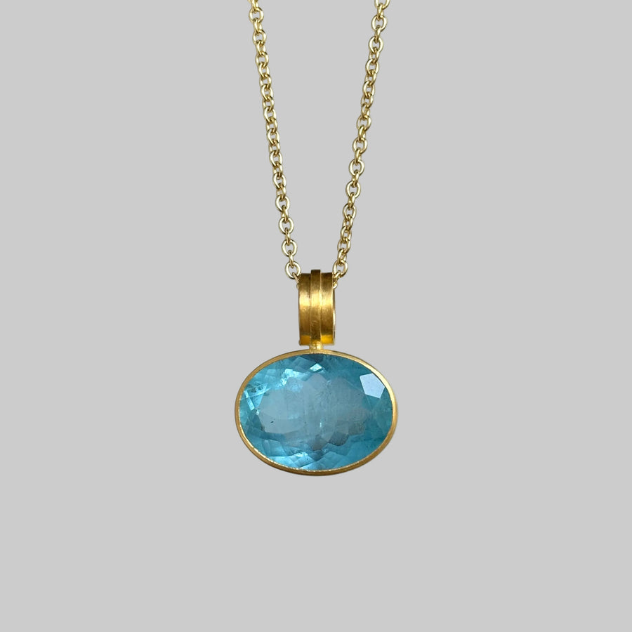 A gold chain necklace with an oval light blue gemstone pendant in a gold bezel setting, shown against a plain light gray background.