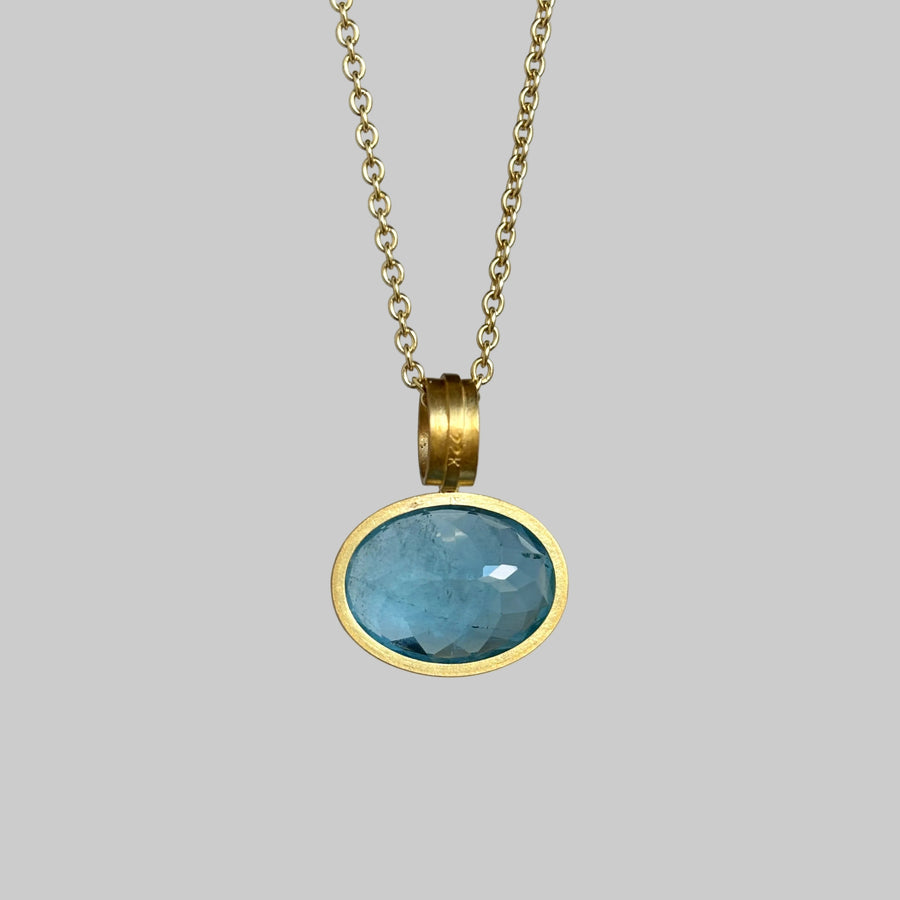 A gold chain necklace with an oval bezel-set light blue gemstone pendant, shown against a plain light gray background.