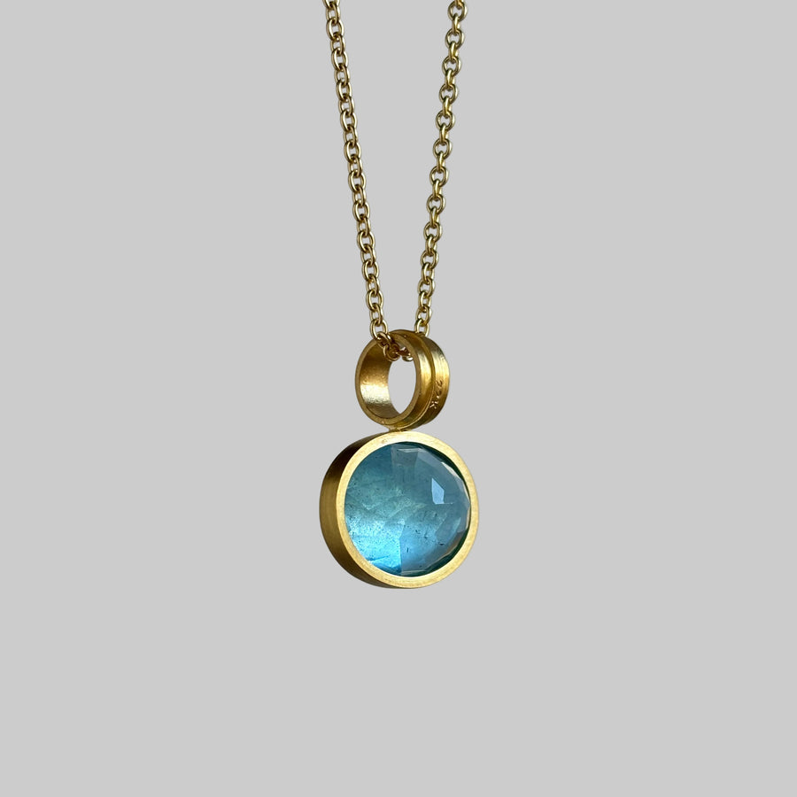 A gold chain necklace featuring a round bezel-set light blue gemstone pendant with a small gold ring spacer above it, shown against a solid gray background.