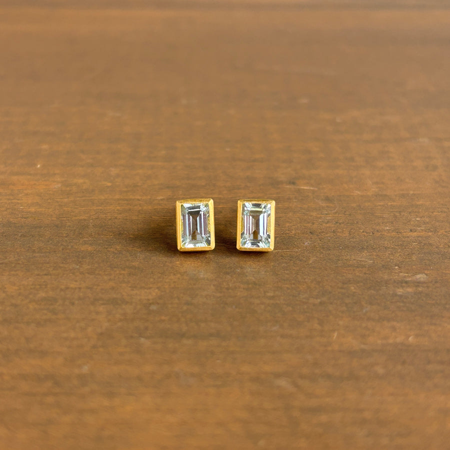 A pair of gold stud earrings with rectangular clear gemstones, shown on a brown wooden surface. 