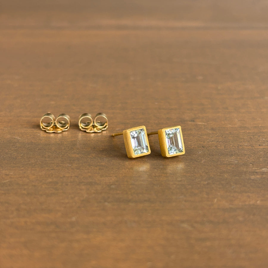 A pair of gold stud earrings with rectangular clear gemstones, shown on a brown wooden surface. One earring is displayed in front in profile, showing the post and back, while the other is turned to show the back post.