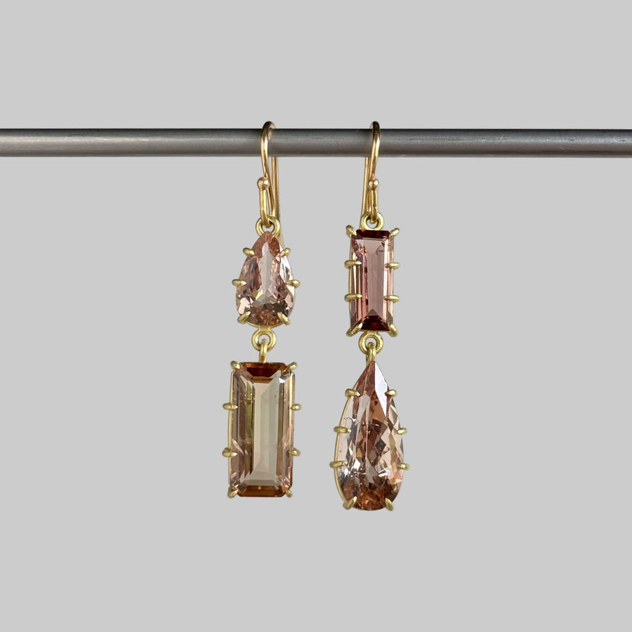 A pair of gold drop earrings with rectangular and teardrop-shaped blush pink gemstones, hanging from a horizontal metal bar against a light gray background.