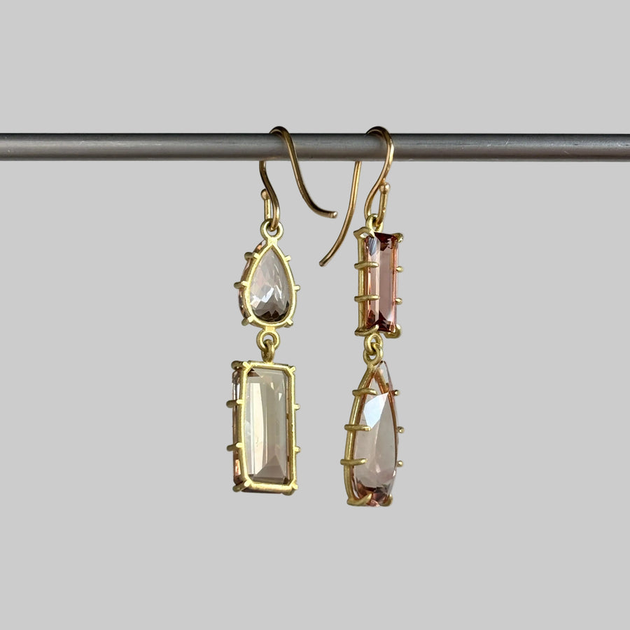 A pair of gold drop earrings with marquise and rectangular clear faceted gemstones, hanging from a dark metal rod against a light gray background.