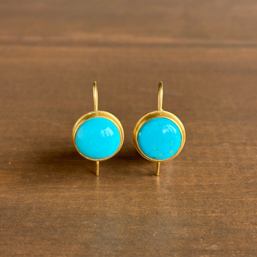 A pair of gold drop earrings with round turquoise cabochon stones, displayed on a dark brown wooden surface.