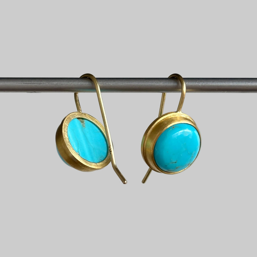 A pair of gold-tone drop earrings with oval turquoise stones, hanging from a metal rod against a light gray background.
