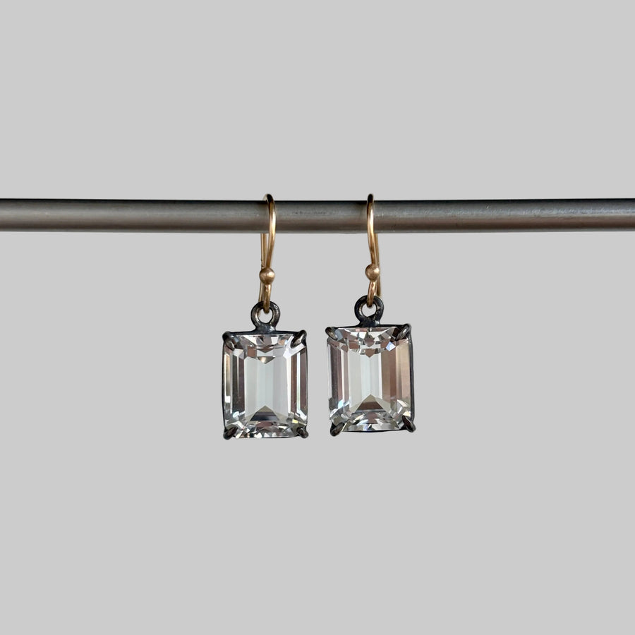 A pair of rectangular clear crystal drop earrings with gold-toned fishhook backings, hanging from a dark horizontal bar against a light gray background.
