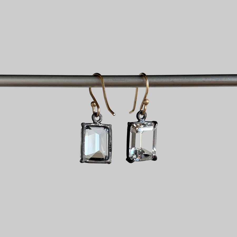 A pair of rectangular clear crystal drop earrings with silver-toned bezel settings and gold-tone fishhook ear wires, hanging from a horizontal metal rod against a light gray background.