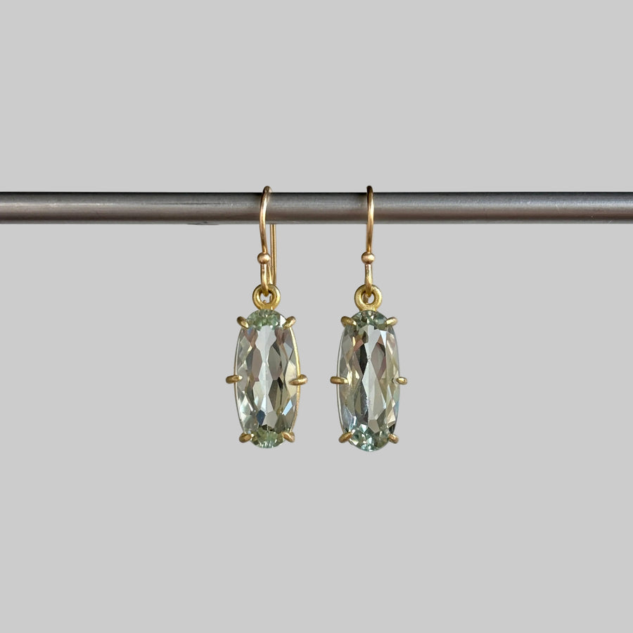 A pair of gold drop earrings with  light green gemstones, hanging from a dark metal rod against a soft light gray background.