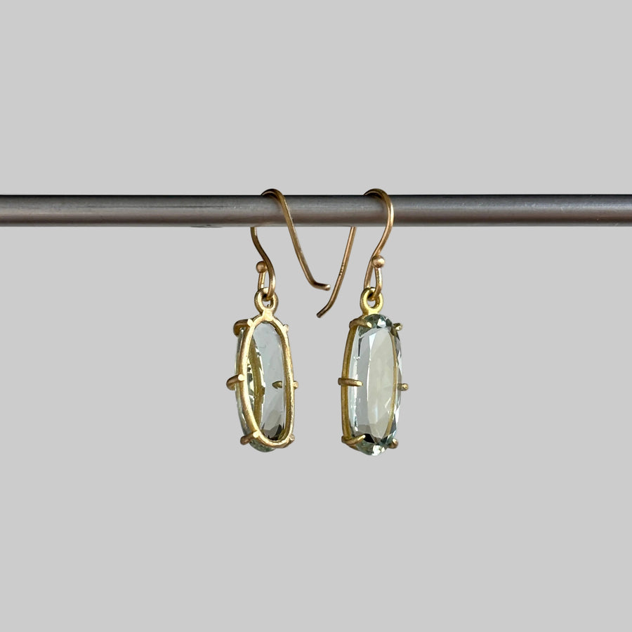 A pair of gold drop earrings with oval clear gemstones, hanging from a dark metal rod against a light gray background.