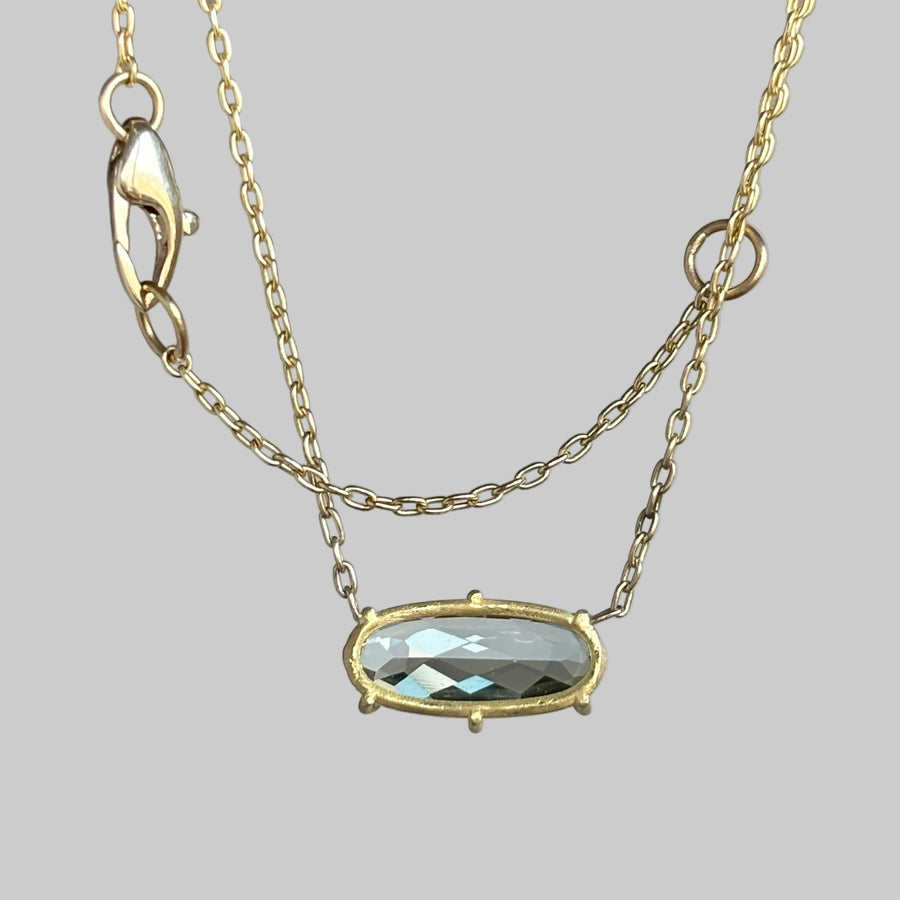 A delicate gold chain necklace featuring a small oval bezel-set faceted gray gemstone pendant, displayed against a soft light gray background.
