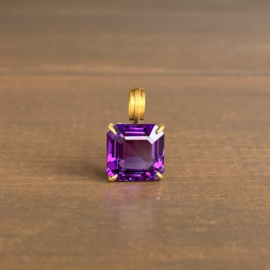 A square-cut purple gemstone pendant in a gold setting, resting on a brown wooden surface.