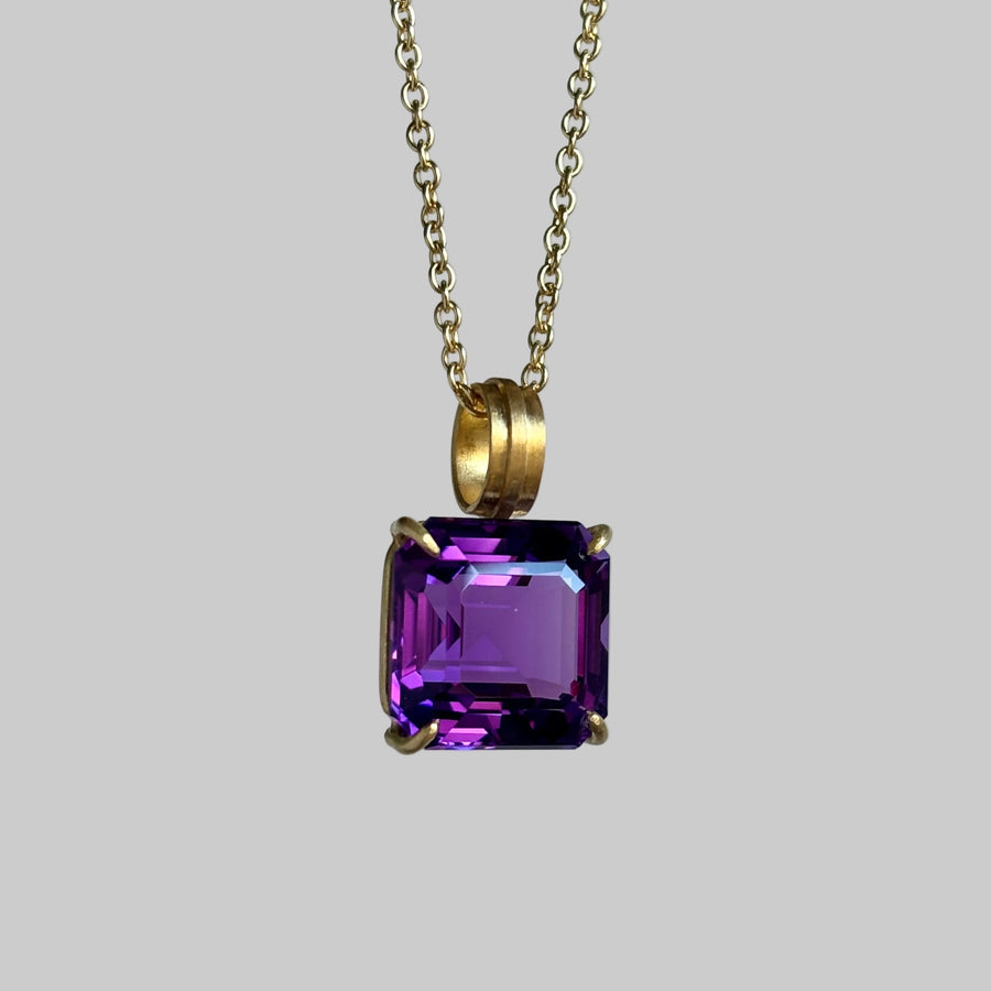 A gold chain necklace with a square-cut purple gemstone pendant in a gold setting, shown against a plain light gray background.