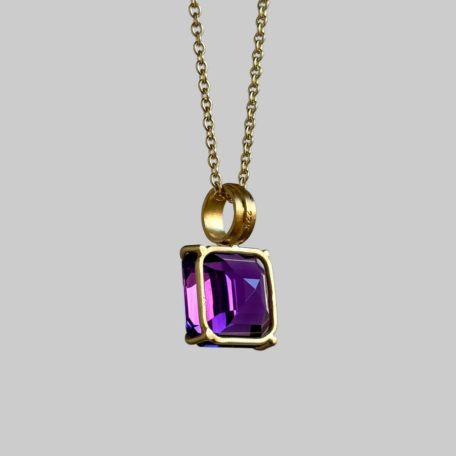 A gold chain necklace with a square bezel-set purple crystal pendant, shown against a solid gray background.