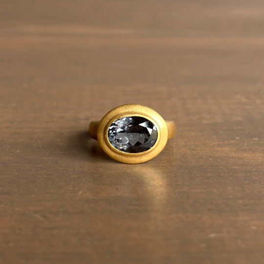 An oval-shaped ring with a grey tourmaline centerpiece, set in a yellow gold bezel-style mounting, placed on a wooden surface. Rosanne Pugliese Greek-Inspired Grey Tourmaline Ring