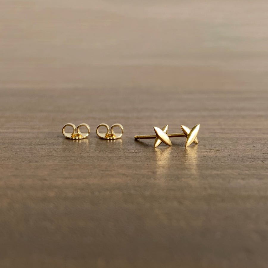 A pair of small star-shaped cross stud earrings in 18k yellow gold, placed on a wooden surface.