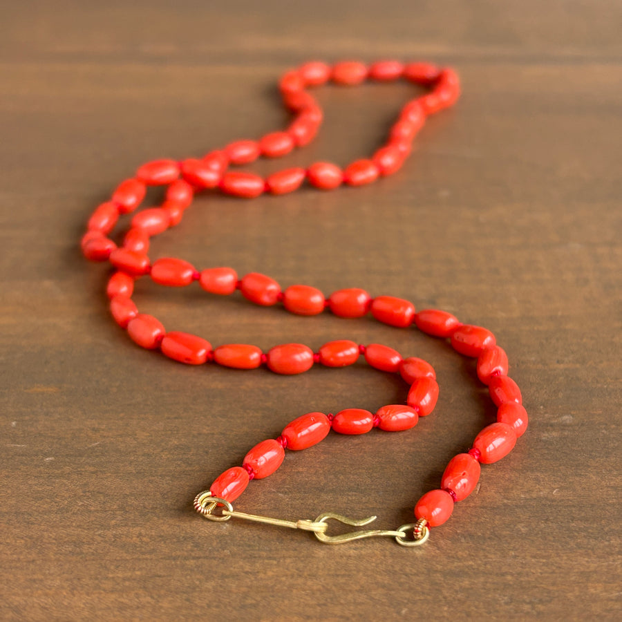 A coral red beaded necklace with a gold safety-pin style clasp, displayed on a dark brown wooden surface.