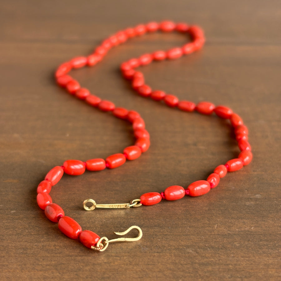 A red beaded necklace with elongated oval beads and gold-toned hardware, displayed on a dark brown wooden surface.