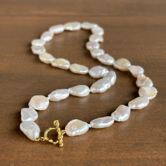 A baroque pearl necklace with a gold toggle clasp, displayed on a dark brown wooden surface.