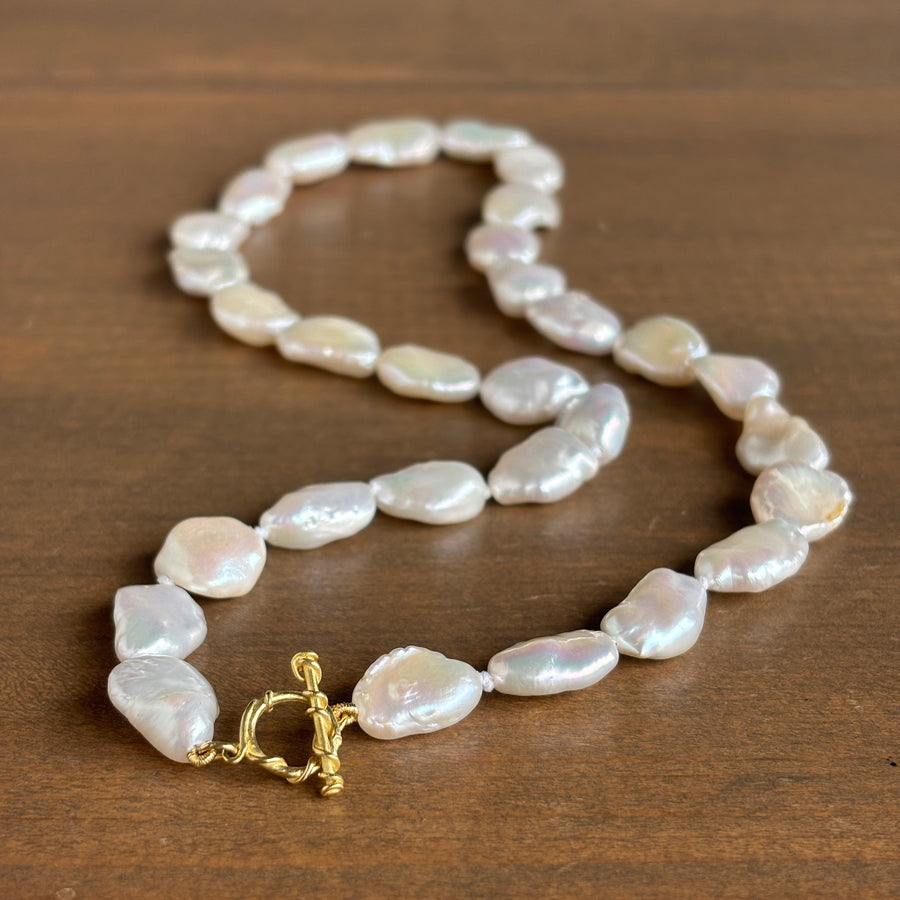 A baroque pearl necklace with a gold toggle clasp, displayed on a dark brown wooden surface.