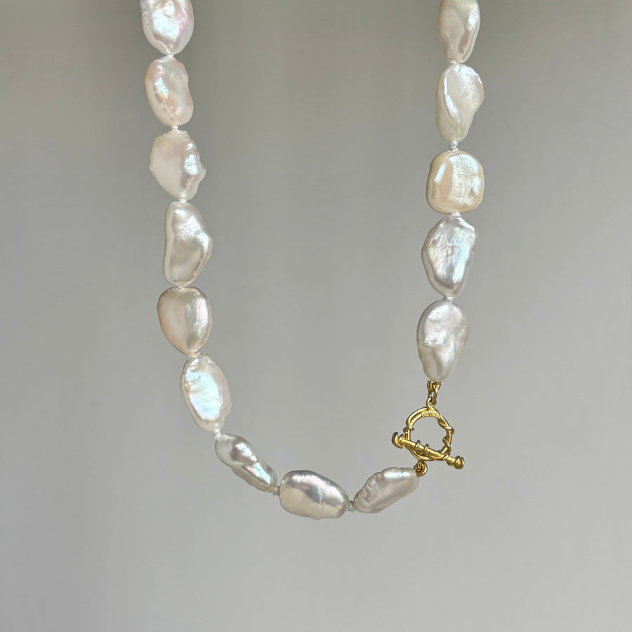 A baroque pearl necklace with irregular, lustrous white pearls and a small gold toggle clasp, photographed against a neutral gray background.