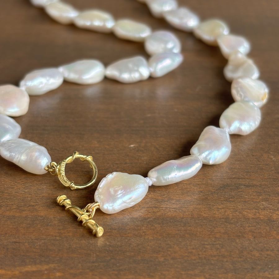 A close-up of a baroque pearl necklace with a gold toggle clasp, resting on a dark brown wooden surface.