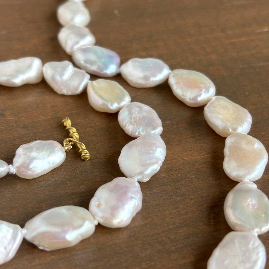 A close-up of a baroque pearl necklace with irregular, lustrous white pearls and a small gold clasp, laid on a dark brown wooden surface.