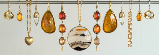 A collection of gold jewelry, including necklaces, earrings, and pendants, hanging from a metal rod against a plain light gray background. The jewelry features amber, orange, red, and clear gemstones in various shapes and sizes.